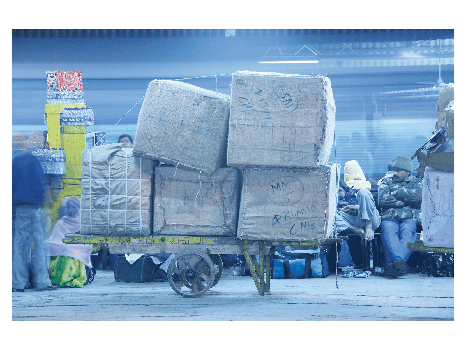 Fine art photography by André Wagner: Large bundles secured with rope are stacked on a low trolley — Am hauptbahnhof in neu-dehli, Indien.