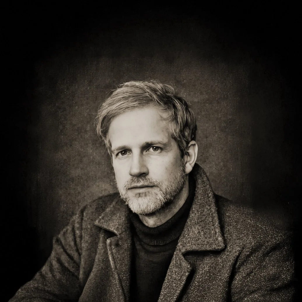 Portrait en noir et blanc de l’artiste photographe André Wagner en manteau sombre sur fond sombre.