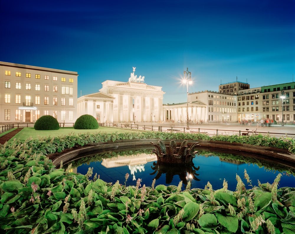 Fine art photography by André Wagner: A round pool surrounded by dense planting occupies the foreground — Brandenburg Gate, Deutschland.
