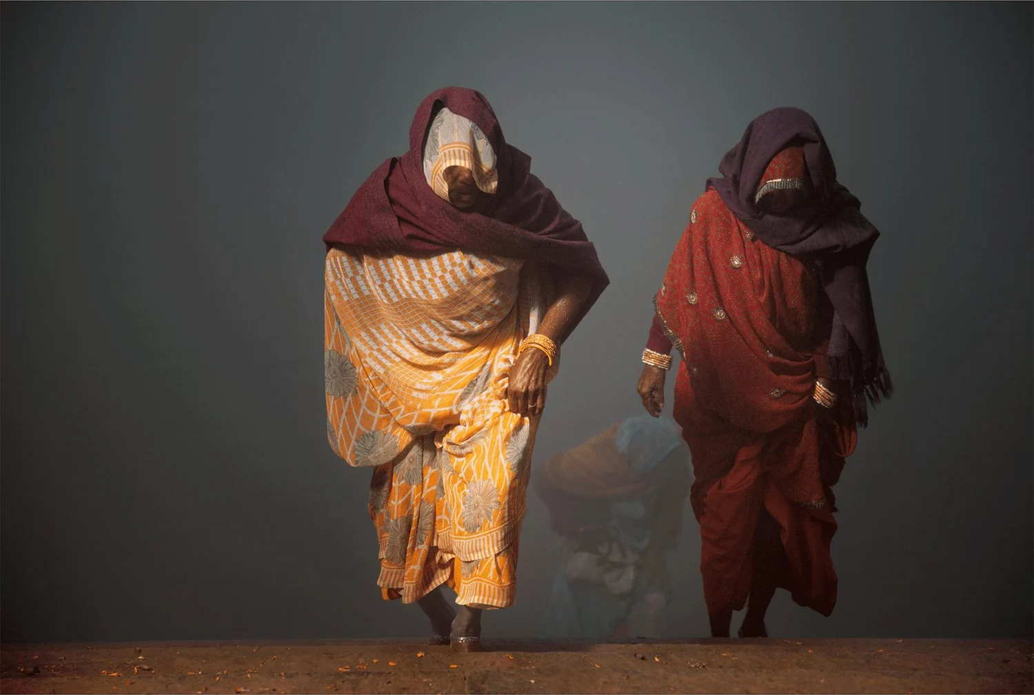 Fotokunst von André Wagner: Zwei Frauen treten deutlich aus dem grauen Nebel hervor, während hinter ihnen eine weitere Gestalt nur schwach erkennbar bleibt — Coming back from yamuna river, Indien.