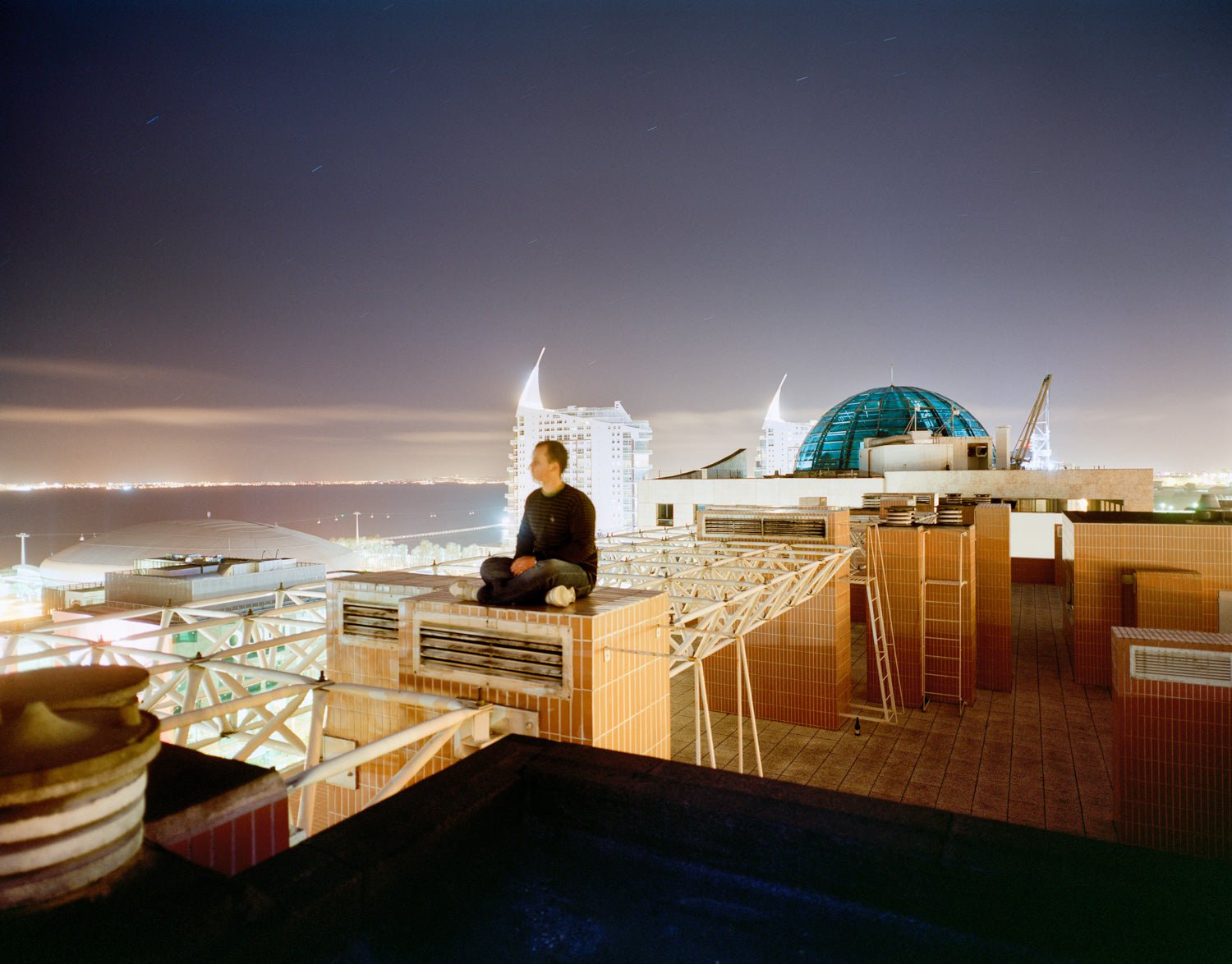 Photographie d’art d’André Wagner: Une personne est assise, les jambes croisées, sur une structure basse du toit — East Wind, Portugal.