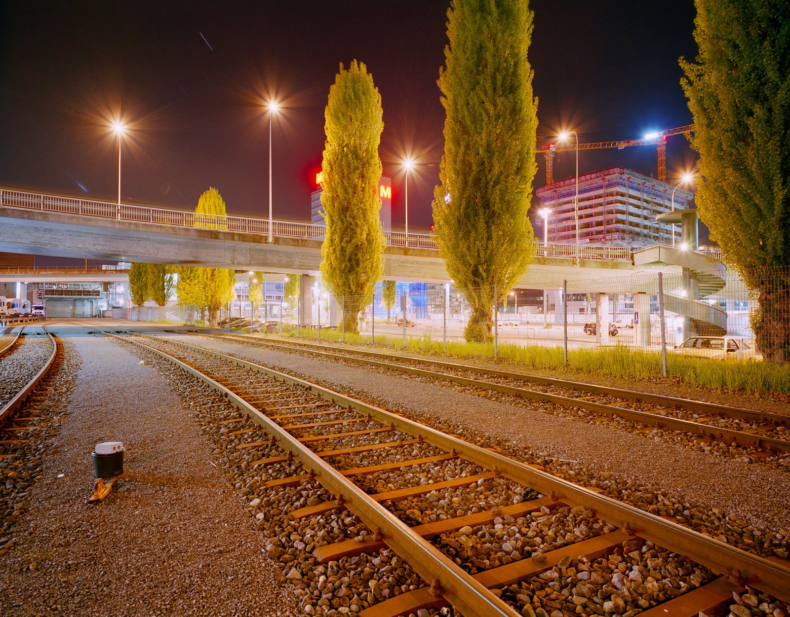 Photographie d’art d’André Wagner: Des rails décrivent une courbe entre le ballast et des arbres élancés vers un paysage urbain éclairé — Five Stars.