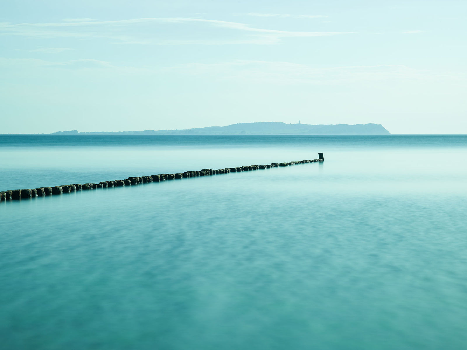 Photographie d’art d’André Wagner: Une étroite rangée de pieux sombres s’avance depuis le bord gauche loin dans l’eau claire — Hiddensee 1, Deutschland.