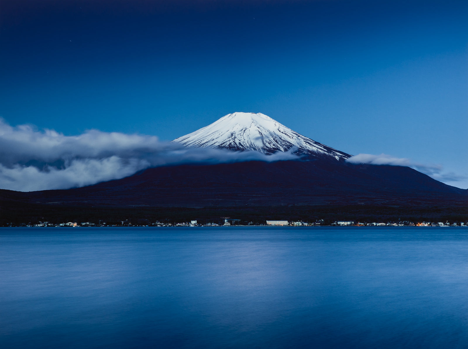 Photographie d’art d’André Wagner: Une montagne enneigée se détache en une forme claire, presque triangulaire, au-dessus de pentes sombres — Mt. fuji at night, Japan.