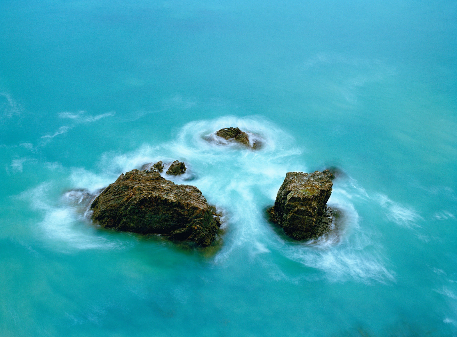 Photographie d’art d’André Wagner: Plusieurs rochers sombres se trouvent dans une étendue d’eau turquoise qui remplit presque l’image — Ocean, Neuseeland.