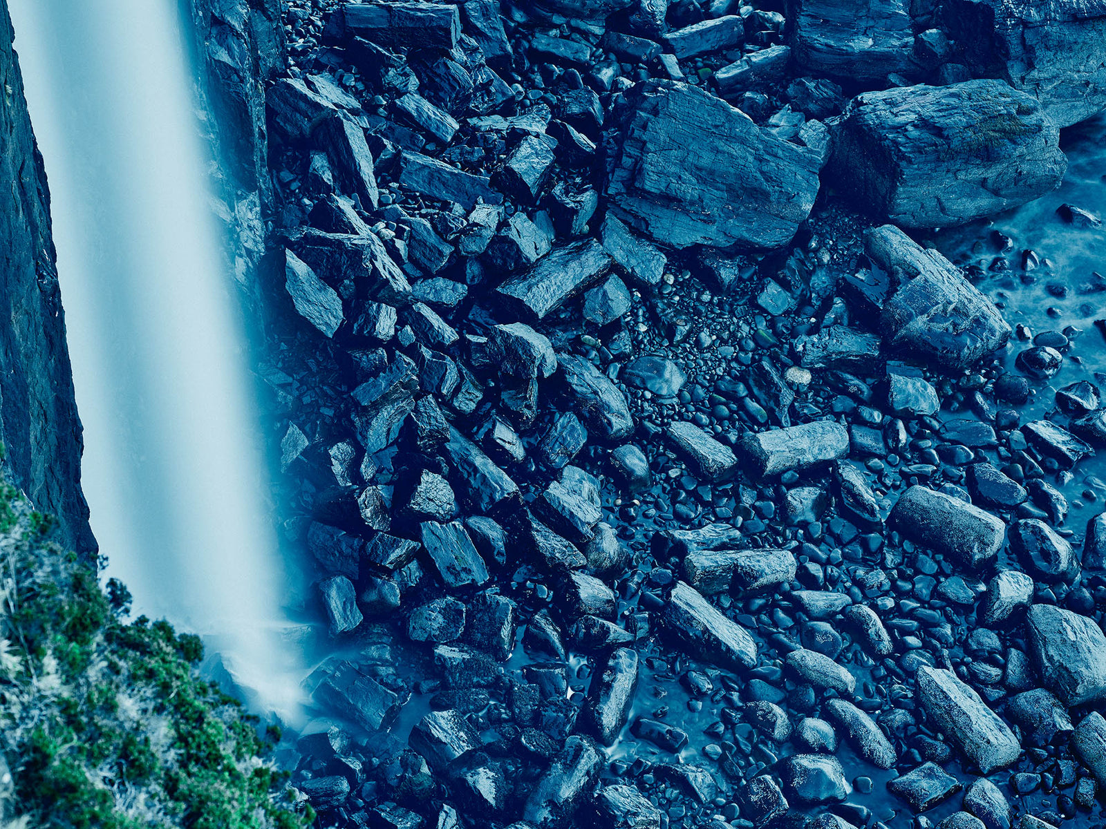 Photographie d’art d’André Wagner: Une cascade claire descend en une bande verticale presque continue le long d’une paroi rocheuse sombre — Ocean rock and waterfall, Schottland.