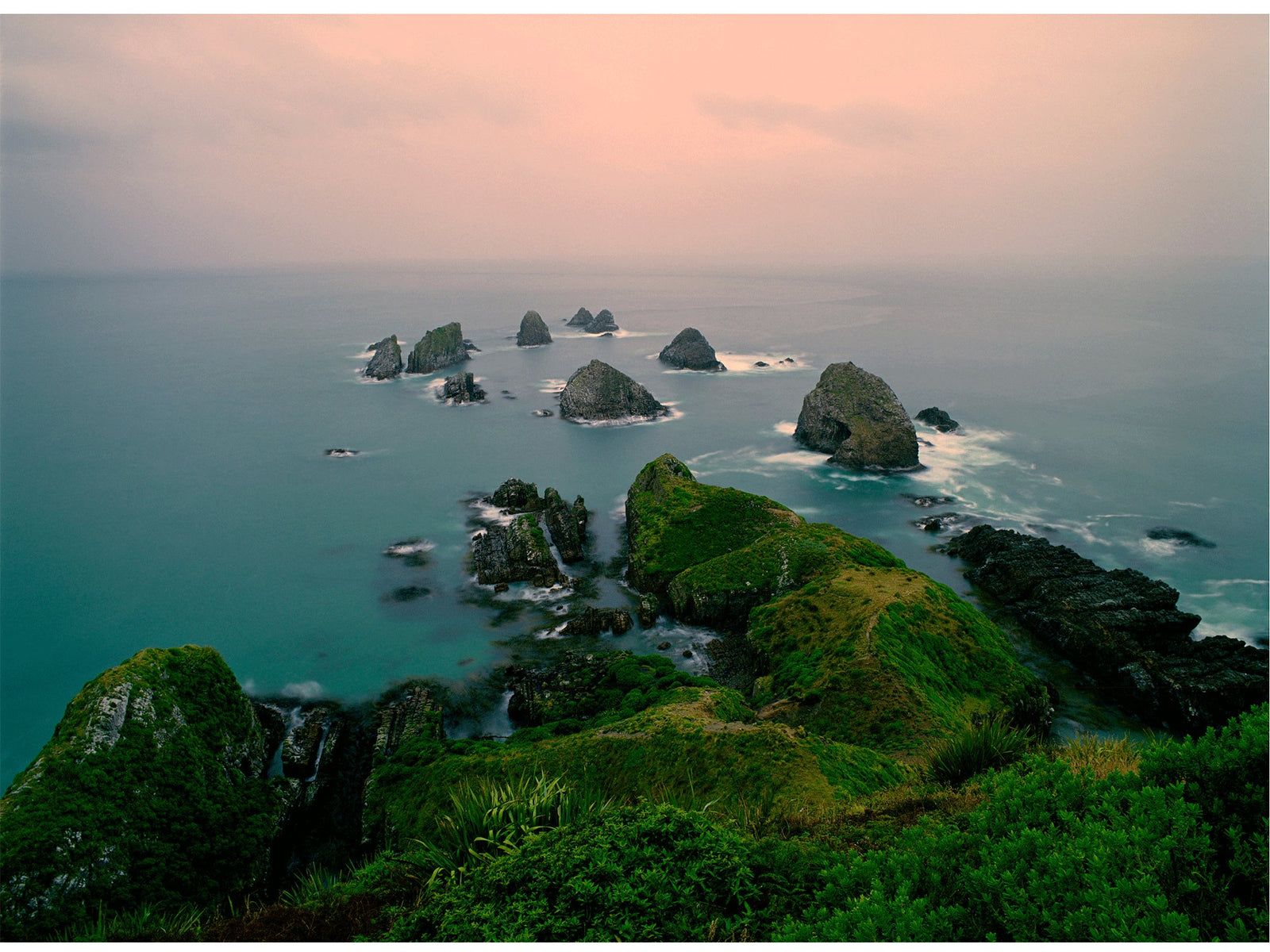 Photographie d’art d’André Wagner: Depuis une bordure côtière végétalisée, une succession de rochers sombres s’avance dans la mer — Ocean rocks, Neuseeland.