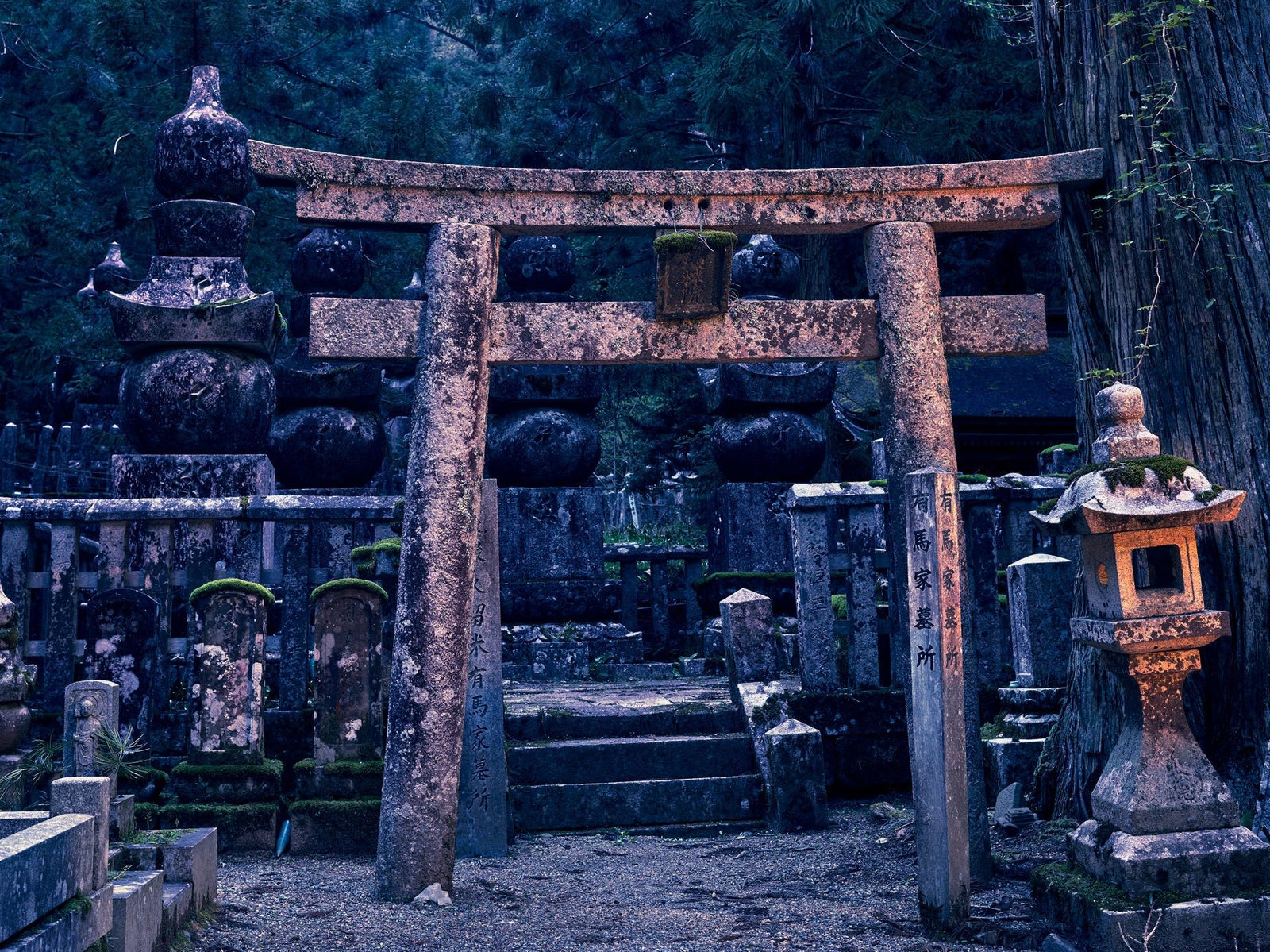 Fine art photography by André Wagner: A stone gate stands frontally among tall trees and smaller stone forms — Oku-No-In Twillight, Japan.
