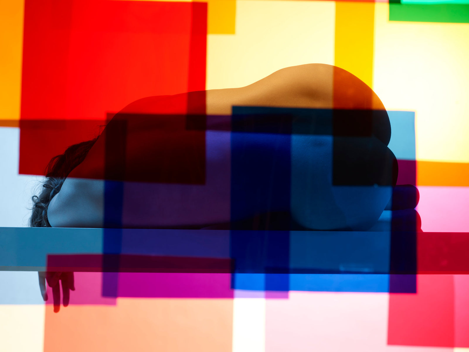 Photographie d’art d’André Wagner: Le corps, étroitement replié, forme une courbe sombre et fermée devant des surfaces colorées claires — Paula II.