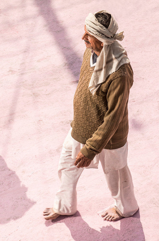 Photographie d’art d’André Wagner: Un homme coiffé d’un foulard clair, vêtu d’un haut brun et d’un pantalon blanc, marche sur un sol rose largement dégagé — Pilgrim on Purple Dust 4, Indien.