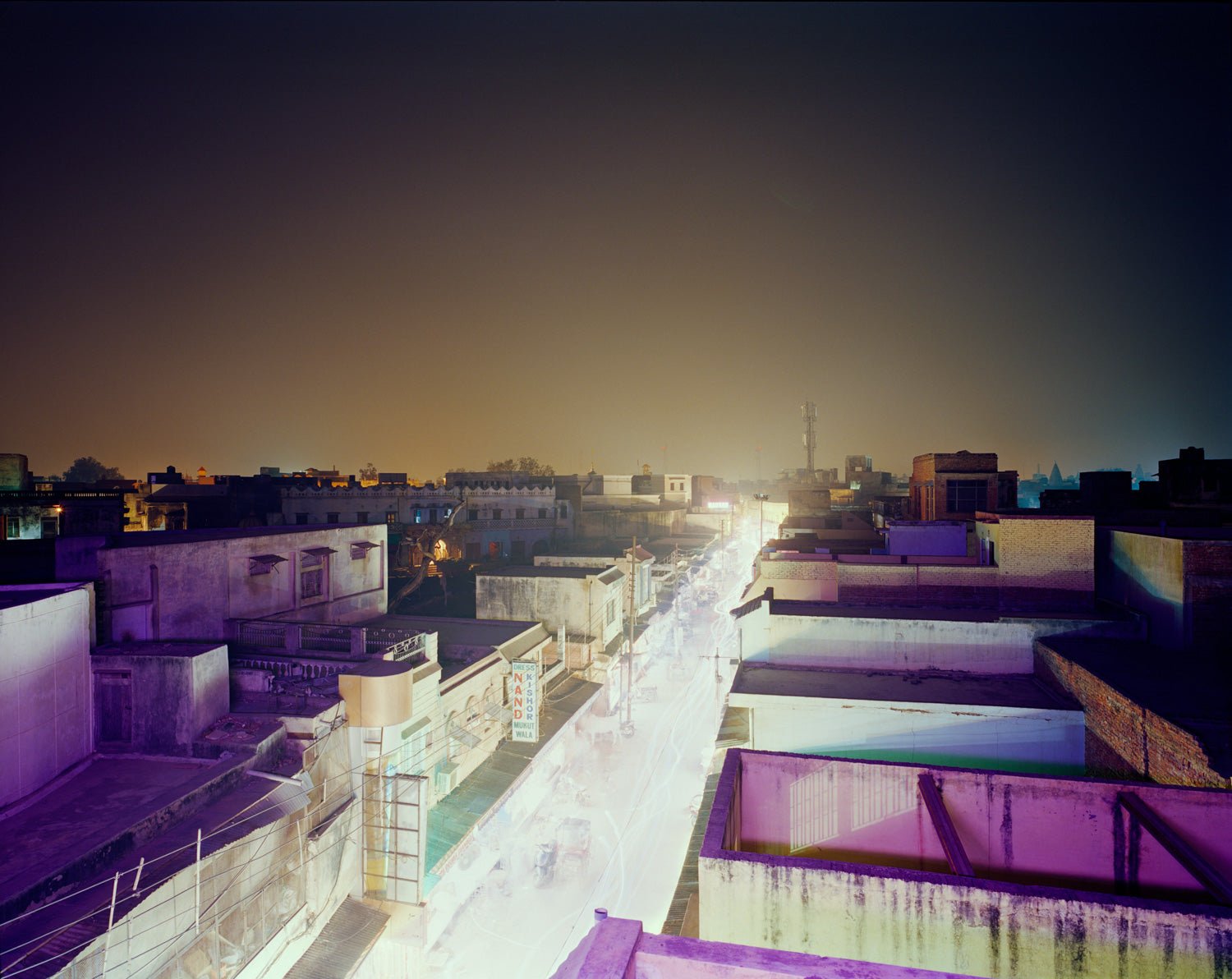 Fine art photography by André Wagner: A brightly lit street runs straight between low roofs — Shopping Street, Indien.