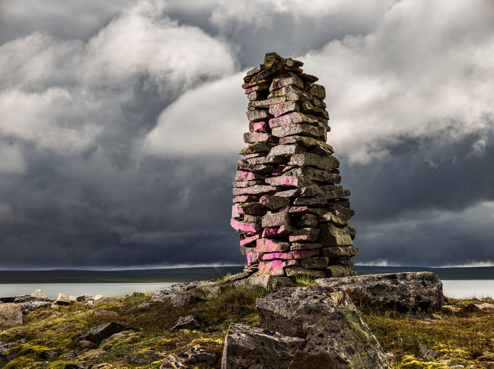 Photographie d’art d’André Wagner: Une haute colonne de pierres étroite se dresse sur une roche rugueuse, en partie recouverte de vert — Stoneman 1, Island.