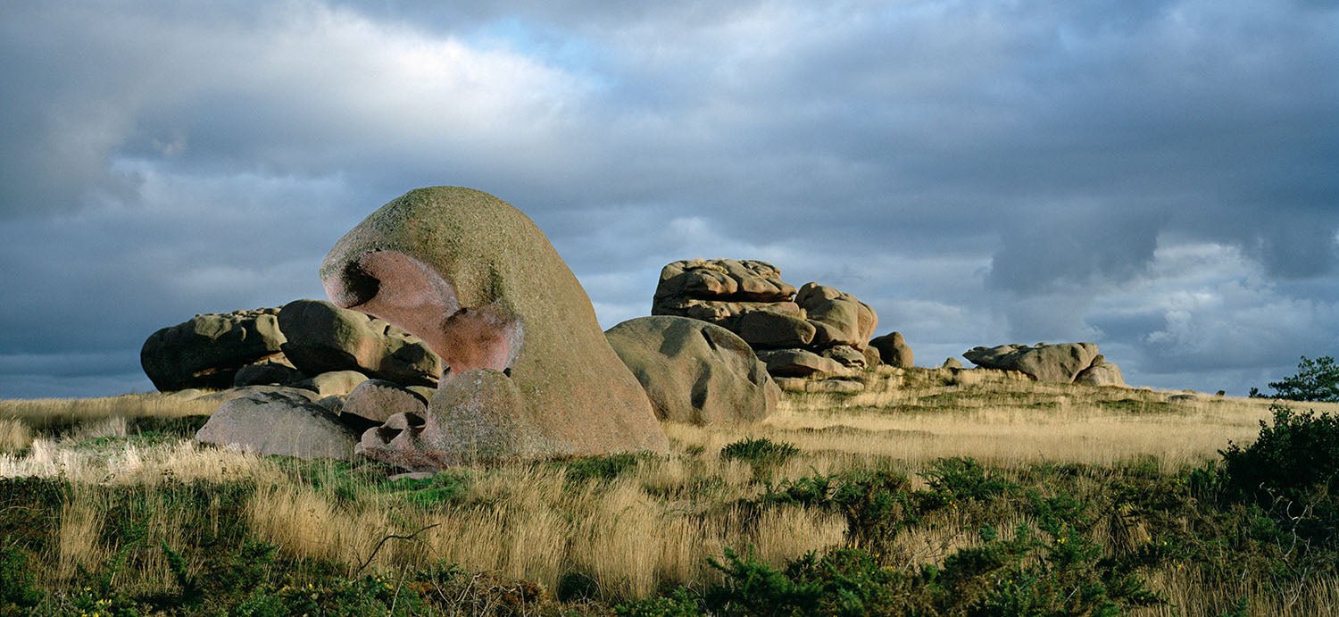 Photographie d’art d’André Wagner: Un grand rocher arrondi se dresse parmi de plus petites pierres dans une étendue d’herbe plate — Story of wind 2, Frankreich.