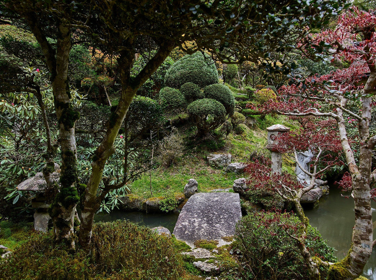 Fine art photography by André Wagner: A small stone bridge crosses dark water between trees — Temple in oku-no, Japan.