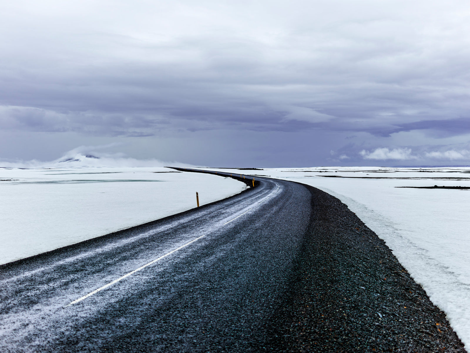 Photographie d’art d’André Wagner: Une route sombre décrit une courbe depuis le bord inférieur droit vers une vaste plaine blanche — Way To The End, Island.