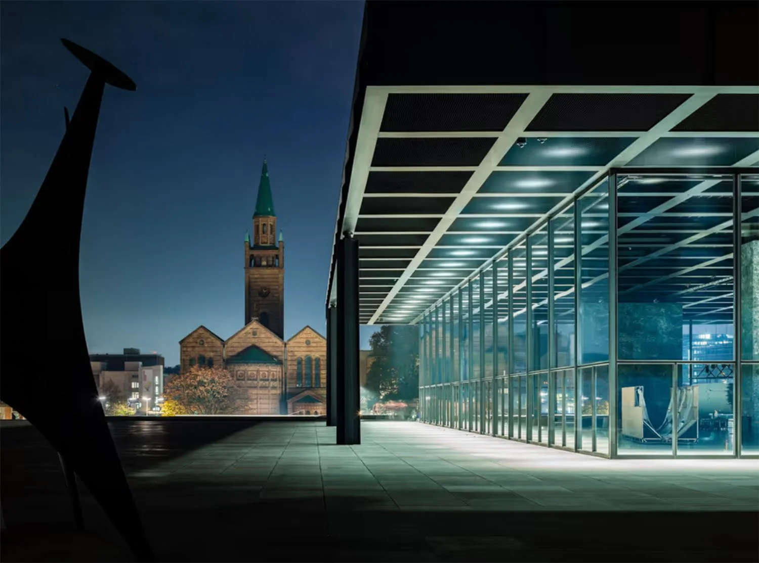 Moderne Glasfassade eines Geb&auml;udes bei Nacht mit Blick auf eine historische Kirche im Hintergrund.