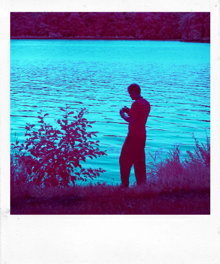 Photographie infrarouge d’une personne debout au bord d’un lac, dans des tons magenta et turquoise.