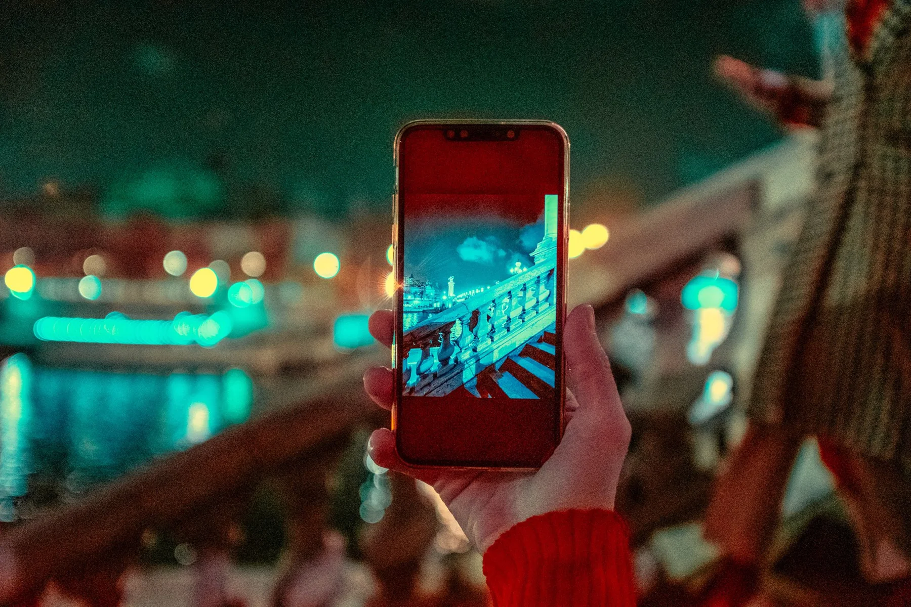 Ein Smartphone am Wasser vor einer n&auml;chtlich beleuchteten Stadtkulisse.